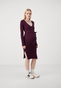 Maternity dress in ribbed dark purple fabric, V-neck, long sleeves, fitted silhouette with side slits, paired with white sneakers.