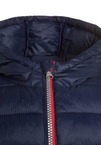 Granatowa kurtka puchowa z kapturem, z srebrnym zamkiem błyskawicznym z przodu oraz czerwonymi akcentami materiałowymi wzdłuż zamka. Błyszcząca, pikowana struktura.