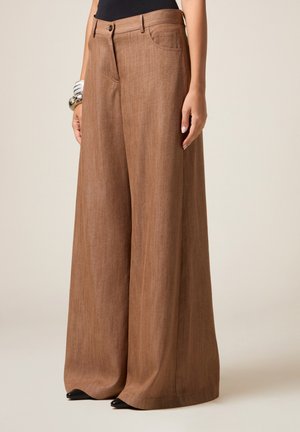 Pantaloni a gamba larga marroni indossati con scarpe nere a punta e bracciali d'argento sul polso sinistro, mostrati dalla vita fino al pavimento.
