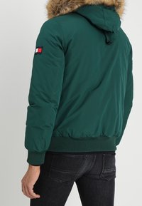 Giacca imbottita verde con cappuccio in pelliccia sintetica, polsini a coste e una toppa con logo sul braccio sinistro. Il retro presenta una superficie liscia con un design aderente.