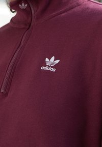 Sudadera con cremallera color marrón burdeos en tejido suave, con un logo blanco de Adidas en el lado izquierdo del pecho y un cuello con cremallera corta.