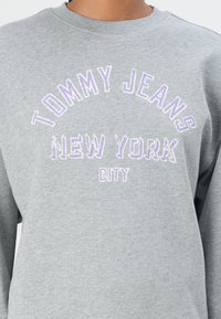 Felpa grigia realizzata in morbido tessuto, con bordature ricamate con il testo "TOMMY JEANS NEW YORK CITY" in viola e bianco, e scollo a giro.