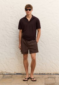 Korte mouwen, donkergroene poloshirt gecombineerd met bijpassende shorts. Zwarte slippers. Staand tegen een gestructureerde lichtgekleurde muur.