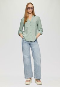 Lichtgroen overhemd met lange mouwen, gecombineerd met wijd uitlopende lichtblauwe jeans en platformsandalen met opvallende banden.