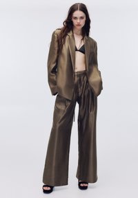 Olivegrønt satinblazer og buksedragt med vide ben og blank finish; har en kontrasterende sort bh under. Sorte plateau-sandaler inkluderet.