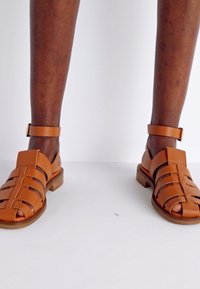 Braune Leder-Sandalen mit gewebtem Design, Knöchelriemen und niedrigem Holzabsatz. Offene Zehen und robuste Verarbeitung.