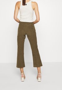 Femme de dos portant un pantalon court en velours côtelé olive, un haut sans manches blanc et des sandales à talons carrés beiges sur fond blanc.