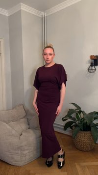 O rochie lungă burgundy, mulată, cu mâneci scurte tip aripă de liliac, dintr-un material texturat, asortată cu pantofi negri cu curele și catarame, pe un fundal neutru.