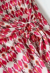 Tissu avec un motif abstrait rouge et orange rassemblé et enfilé à travers un anneau métallique argenté, créant des plis et une texture sur un fond clair.