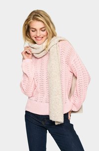 Maglione rosa chiaro a maglia con motivo a punto aperto, abbinato a una sciarpa crema con trama. Il modello indossa jeans scuri.
