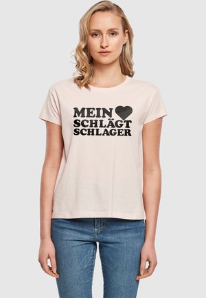 Heled roosa puuvillane T-särk lühikeste varrukatega, ümar kaelus ja must trükk "MEIN ♥ SCHLÄGT SCHLAGER" silmapaistvalt esiküljel.
