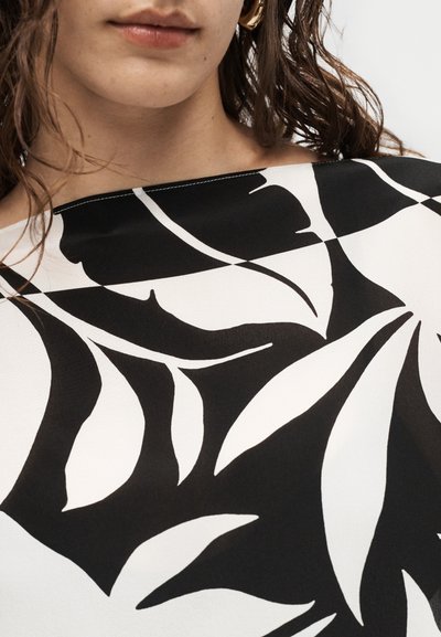 Νέα γυναίκα που φοράει λευκό μπλουζάκι με μεγάλα μαύρα αφηρημένα σχέδια φύλλων και χρυσούς κρίκους στα αυτιά, εμφανιζόμενη από το πηγούνι μέχρι τους ώμους.