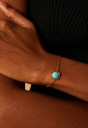 Gouden kettingarmband met een ronde turquoise steen in een hexagonale zetting, gedragen op een bruine huidskleur. Geweven zwarte achtergrond zichtbaar.