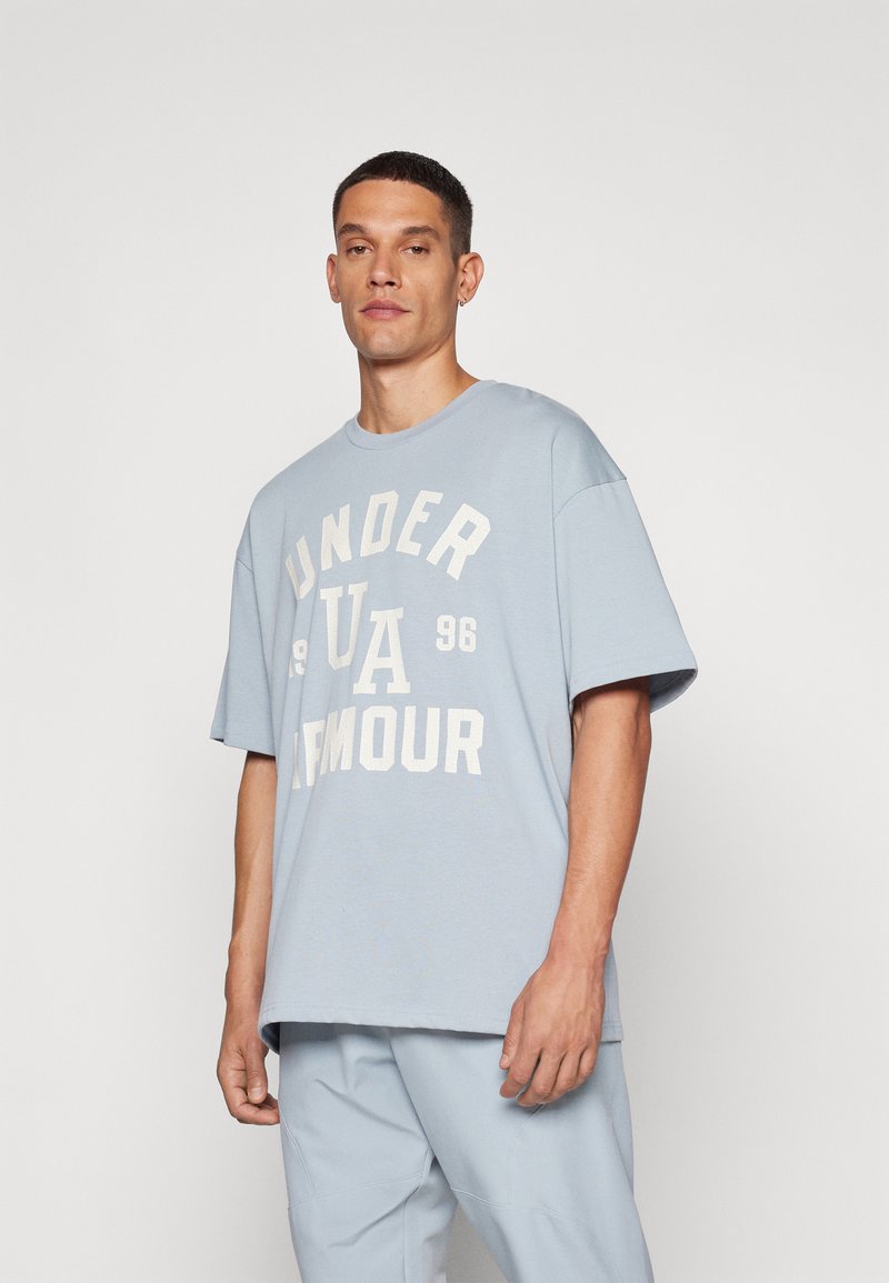 Camiseta de algodón de color azul claro con cuello redondo, que presenta un texto blanco en negrita que dice "UNDER ARMOUR 1996" en la parte frontal. Mangas cortas.