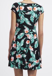 Kobieta w czarnej sukience z krótkim rękawem z zielonymi liśćmi palmowymi i pomarańczowymi wzorami kwiatowymi, pokazana od tyłu.
