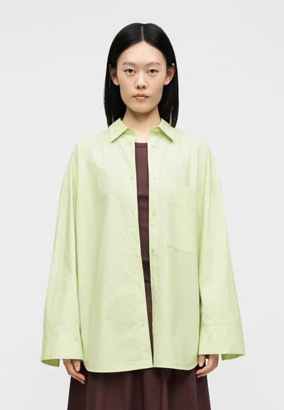 Jeune femme aux cheveux noirs porte une chemise oversize vert clair boutonnée sur une robe marron, regardant vers l'avant sur un fond uni.