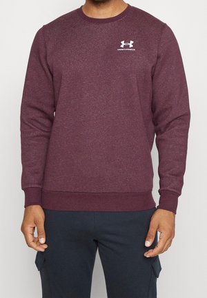 Mann trägt ein burgunderfarbenes Under Armour Sweatshirt mit Logo auf der Brust und dunkelblaue Cargohosen, steht vor einem schlichten Hintergrund.