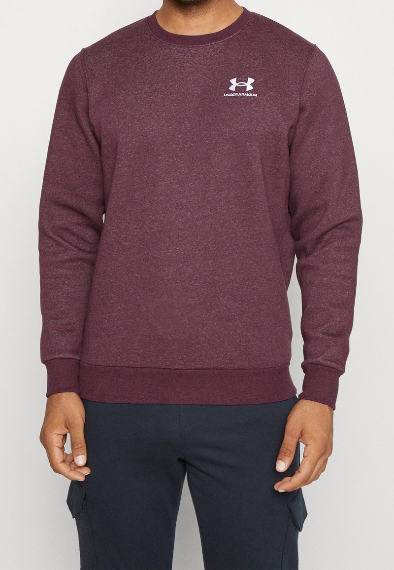 Homme portant un sweat-shirt Under Armour bordeaux avec un logo sur la poitrine et un pantalon cargo bleu foncé, debout devant un fond uni.