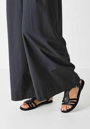 Black sandals with multiple straps and decorative studs, flat sole, and ankle buckle. Paired with loose-fitting black pants.