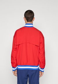 Red bomber jacket with a blue and white striped collar and cuffs. Features a vent detail at the back. Smooth texture and relaxed fit.