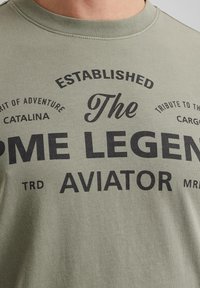 Camiseta de algodón verde oliva con texto gráfico en negro que incluye frases como "Establecido," "El Aviador PME Legend," y varios acentos.