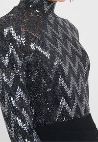 Svart paljett-topp med hög hals, med ett zigzag-mönster i silver. Tyget är slätt med en glittrande textur.