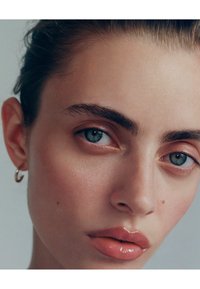 Primer plano de un rostro con piel clara, ojos azules, cejas definidas y labios ligeramente entreabiertos con un acabado brillante en tono rosa. Se ve un pendiente de aro dorado.