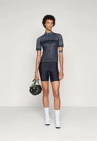 Mustas tsüklisärk ja lühikesed püksid kandv inimene hoiab musta jalgrattakiivrit, jalas valged sokid ja valged tsüklijalatsid, seistes neutraalse tausta ees.