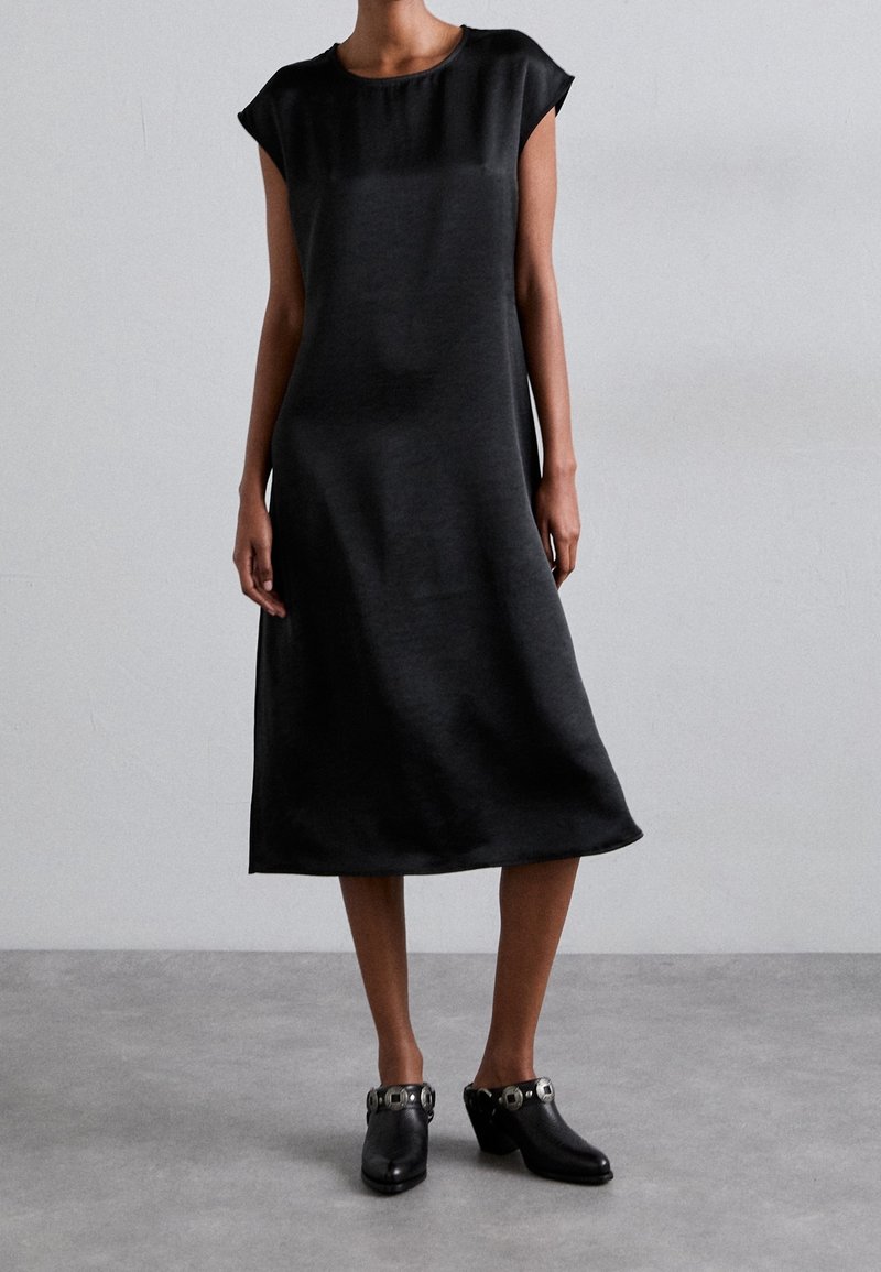 Femme portant une robe midi noire à manches courtes et des mules noires à talons avec des détails en boucles en argent, se tenant devant un fond gris uni.