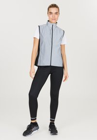Gilet smanicato grigio chiaro con colletto alto, zip frontale e pannelli laterali neri, abbinato a leggins neri aderenti e scarpe da corsa nere.