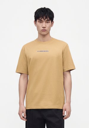 Homme aux cheveux courts noirs portant un t-shirt beige J.Lindeberg et un pantalon noir, debout devant un fond blanc uni.