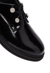 Schwarzer Lacklederschuh mit runder Spitze, ausgestattet mit schwarzen Schnürsenkeln und silbernen Plattenakzenten nahe den Schnürsenkeln, ergänzt durch eine Gummisohle.