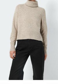 Pull à col roulé beige en tricot avec poignets et ourlet côtelés, porté avec un pantalon noir taille haute sur une personne debout.