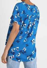 Blue floral short-sleeve top featuring a round neckline and back pleat detail, with white and dark blue flower patterns. Made from soft fabric.