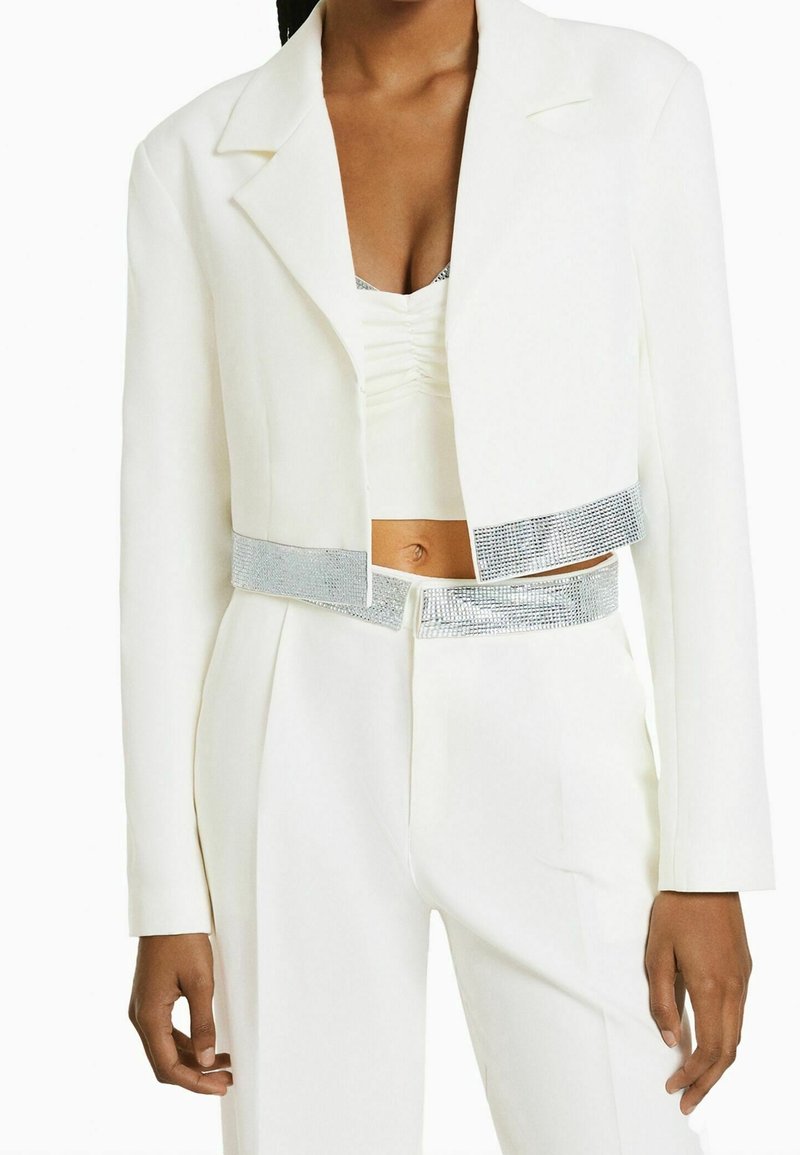 Blazer blanc court et pantalon taille haute assorti avec bordure de strass argentés sur les bords, porté sur un crop top blanc froncé.