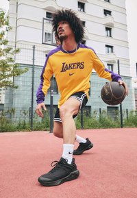 Gul og lilla langærmet Lakers-trøje, sorte shorts med hvide striber og sorte sneakers på en rød multi-sportsbane.