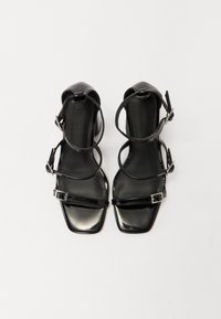 Sandales à talons hauts noires avec un orteil carré, dotées de plusieurs fines lanières et d'une boucle de cheville réglable. Fini en cuir verni brillant.