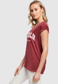 Maronifarvet kortærmet t-shirt med rund hals, der har hvid tekst "BERLIN". Stoffet ser blødt ud og har en afslappet pasform. Båret med hvide bukser.