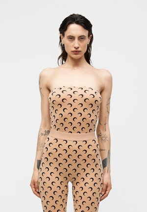 Donna con capelli scuri indossa una tuta beige senza spalline con motivo di lune nere, in piedi su sfondo grigio chiaro.