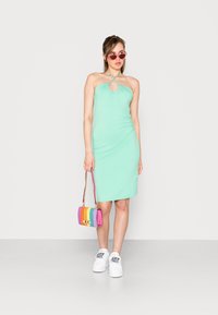 Vestido justo verde menta com декольте halter e detalhe em forma de buraco, combinado com uma bolsa listrada multicolorida e ténis brancos.