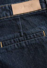 Dark blue denim fabric with back pocket and beige leather patch stitched above, embossed with "JJXX" brand letters.