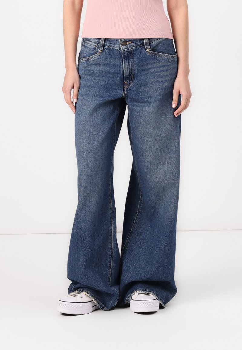 Wijduitlopende jeans in donkerblauwe denim, met een hoge taille, vijf zakken en franjes aan de onderkant, gecombineerd met witte sneakers.