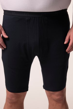 Mann trägt schwarze knielange Shorts und ein weißes, eng anliegendes Hemd, steht mit entspannt an den Seiten hängenden Händen.