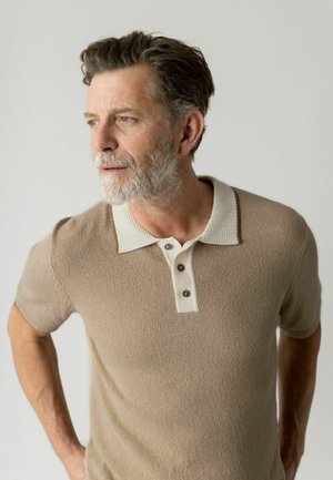 Mand i midalderen med gråt skæg iført beige polo i kortærmet, kigger mod venstre mod en ensfarvet lys baggrund.