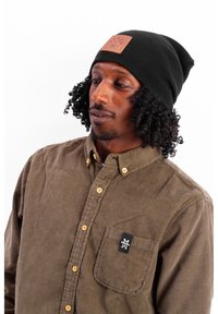 Black knit beanie with a brown leather patch. Brown corduroy button-up shirt with orange buttons and a small black logo on the pocket.