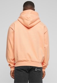Persona che indossa una felpa pesco con cappuccio abbassato e jeans neri con logo bianco ricamato sulla tasca posteriore, di spalle su uno sfondo semplice.