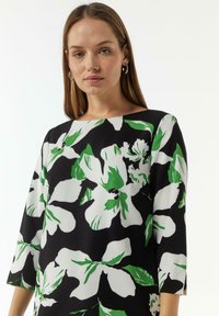 Blumenkleid mit großen weißen und grünen Blumenmustern auf schwarzem Hintergrund, mit quadratischem Ausschnitt und dreiviertel Ärmeln.
