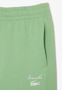 Groene sportbroek van zacht materiaal, met een elastische tailleband, zijzakken en een wit Lacoste-logo met een krokodilafbeelding.