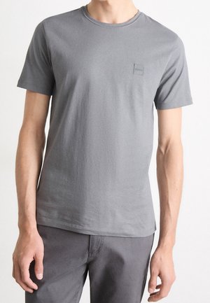 Mann trägt ein schlichtes graues T-Shirt mit kurzen Ärmeln und kleinem Logo auf der Brust sowie passende graue Hose, steht vor weißem Hintergrund.