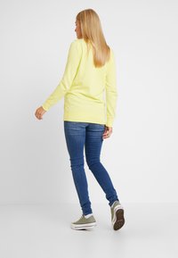 Sweatshirt de manga longa, amarelo brilhante, com decote redondo, combinado com jeans skinny azuis e ténis verde-azeitona. Design simples e casual.
